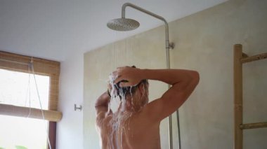 Adam saçlarını şampuanla yıkarken ferahlatıcı duşun tadını çıkarıyor. Zarif bambu mobilyaların olduğu modern banyo. Su taşması günlük kişisel bakım için sakin bir atmosfer yaratır.