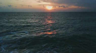 Güneş ufukta batarken, yumuşak okyanus dalgaları kumlu sahile vuruyor. Sıcak altın parıltıyı suya saçıyor. Renkli bulutlu gökyüzünün altında huzurlu ve sakin bir atmosfer yaratıyor..