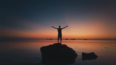 Solan ışıkla çerçevelenmiş gölgeli bir figür. Kaya oluşumunun üzerinde durmakta olan turist silueti, sakin okyanusun üzerinde canlı tropikal günbatımının tadını çıkarıyor, dingin ve ilham verici bir manzara yaratıyor.