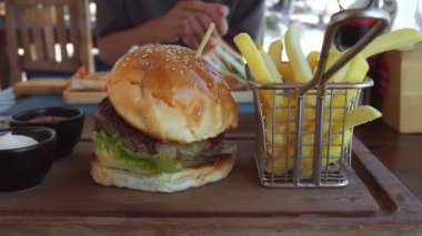 Marullu, domatesli, peynirli ve etli hamburger, metal sepetli patates kızartması ve restoranda ahşap tepside sosla servis edilen arka planda sandviç yiyen bir müşteri.