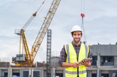 İş, inşaat, endüstri, teknoloji ve insan konsepti - inşaat sahasında tablet bilgisayar kullanırken inşaat alanında çalışan bir grup işçiye kıyasla kasklı neşeli bir inşaatçı