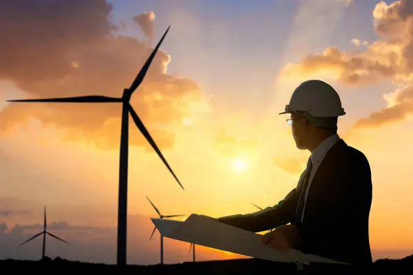 An engineer in hard hat reviews plans against a backdrop of wind turbines and a dramatic sunset.