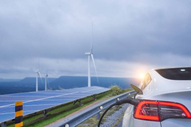 Arka planda rüzgar türbinleri ve güneş panelleri olan şarj istasyonuna takılan elektrikli araba, yenilenebilir enerji kaynaklarını betimliyor