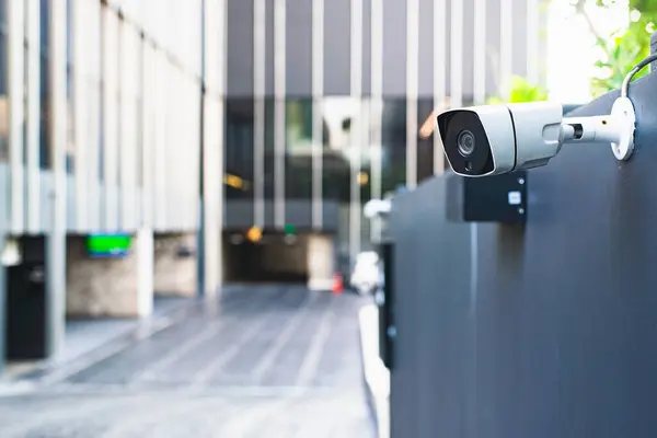 A modern security camera attached to a building\'s exterior wall, overseeing a blurred entrance area