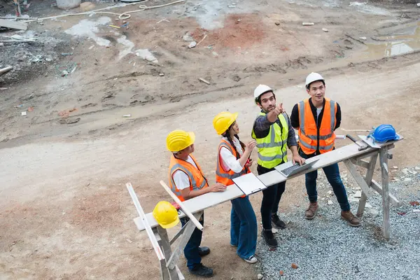 A diverse construction team in safety gear discusses plans over blueprints at a building site