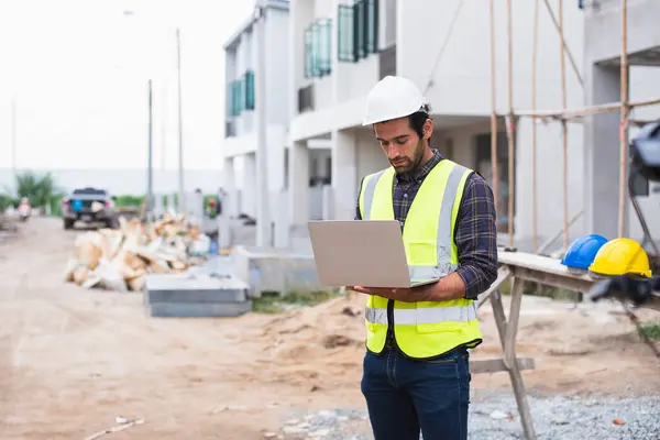 A focused construction supervisor reviews project plans on a laptop at a building site with scattered materials