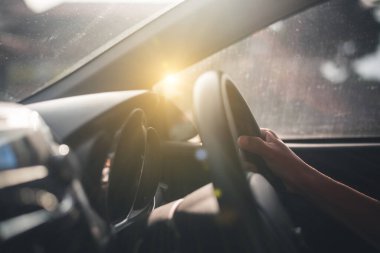 Sürücü ellerinin direksiyondaki yakın görüntüsü. Arka planda güneş batıyor. Yolcu tarafından görülüyor.
