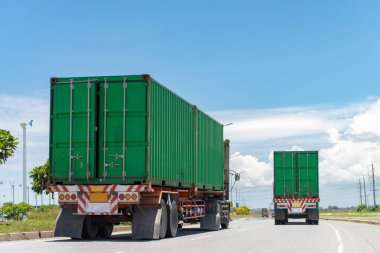 Açık mavi gökyüzünün altında otoyolda giden iki yeşil konteynır kamyonu, ulaşım ve lojistiği vurguluyor..