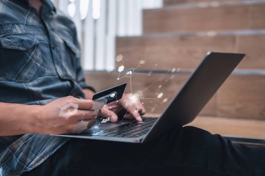 A man in casual attire uses his credit card to shop online, seated with a laptop and digital icons overlay. clipart