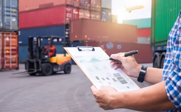 Lojistik müdürü, arka planda forklift ve kargo konteynırları olan bir nakliye deposundaki belgeleri inceliyor..