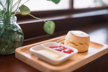 Tahta tepside tereyağı ve çilek reçeli ile servis edilen taze pişmiş bir çörek, saksı bitkisinin yanında..