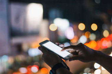 Bulanık şehir manzarasının canlı gece ışıklarıyla arka planında akıllı bir telefonla etkileşime giren ellerin yakın çekimi.