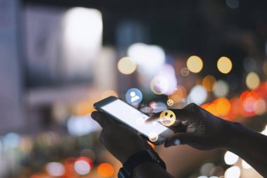 Geceleri sosyal medya simgeleri ve arka planda renkli bokeh ışıkları ile çevrili akıllı bir telefonla iletişim kuran kişi.