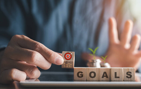 Рука помещает целевой блок рядом со словом "Цели", символизирующим целевую постановку целей, рост и финансовый успех..