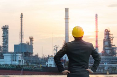 Sarı şapkalı bir mühendisin gün batımında petrol rafinerisi kompleksini gözlemlemesi sanayi gelişimini simgeliyor..