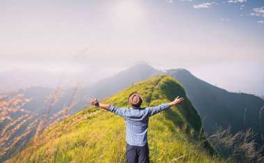 Kolları açık bir adam çimenli bir dağ sırtında duruyor, sabah güneşini kucaklıyor ve uzak tepelerin panoramik manzarasını..