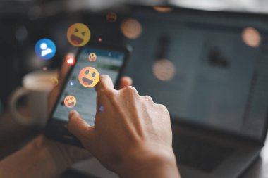 Akıllı bir telefonla etkileşime giren bir elin görüntüsü, sosyal medyanın ilgisini ve etkileşimini sembolize eden renkli yüzen emoji tepkilerini gösterir.