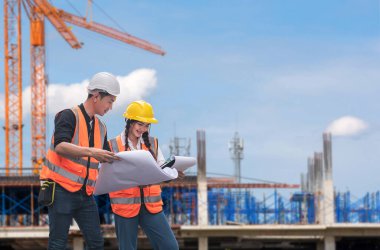 Güvenlik teçhizatı taşıyan iki inşaat işçisi, arka planda vinç ve iskele bulunan bir inşaat alanının planlarını inceliyor..