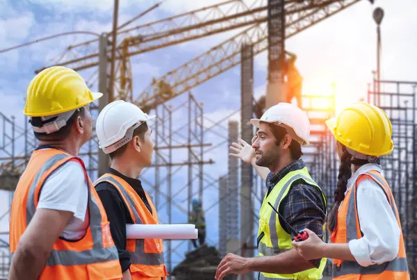 Güvenlik donanımlı çeşitli inşaat işçileri arka planda vinçler olan aktif bir inşaat alanının planlarını tartışıyorlar..