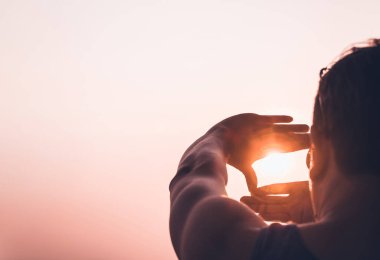 Siluetli bir adam, elleriyle güneşi ılık bir günbatımına karşı çerçeveliyor, görüşü, yaratıcılığı ve olasılıklara bakış açısını simgeliyor..