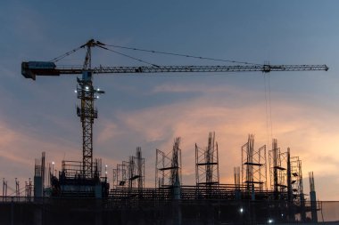 Uzun bir kule vinci ve çelik iskeleti olan bir inşaat alanının silüeti renkli bir günbatımı gökyüzüne karşı kurulmuş..