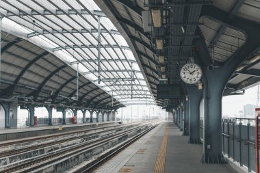 Belirgin bir duvar saati ve tren raylarının yer aldığı eğri büğrü çelik çatı altındaki temiz ve boş bir demiryolu platformunun geniş açılı görüntüsü.