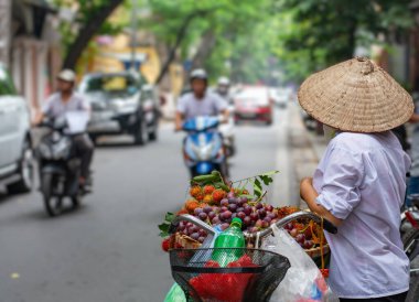 Konik şapka takan geleneksel bir Vietnamlı sokak satıcısı, şehir caddesinde bir bisikletten taze tropikal meyveler satıyor. Sahne, Güneydoğu Asya 'daki günlük yaşam ve canlı sokak kültürünü yansıtıyor..