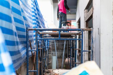 İşçi inşaat iskelesinde dikilirken bir ev duvarını yeniliyor, mavi güvenlik brandaları çalışma alanını kaplıyor..