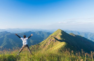 Çimenli bir dağın zirvesinde kolları açık atlayan bir adam, açık mavi gökyüzünün altında manzaralı dağ sıralarıyla özgürlüğü ve macerayı kutluyor..