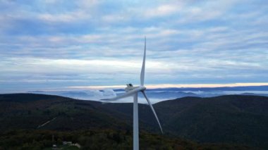 Gün doğumunda dağlarda rüzgar türbinleri ve vadide yoğun sabah sisi. Rüzgar çiftliği yeşil enerji üretiyor.