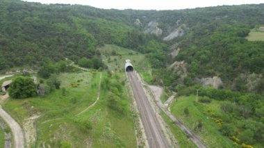 Yüksek hızlı bir yolcu treninin dağın eteklerindeki tünelden çıkarken panoramik drone görüntüsü. Güney Fransa 'daki dağlara tepeden bakan