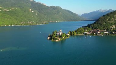 Gün batımında Annecy Gölü 'ndeki Chateau de Duingt' in insansız hava görüntüsü, yemyeşil tepeler ve dağlarla çevrili sakin dağlık sularda süzülen teknelerle. Sinematik altın saat aydınlatması yaratır