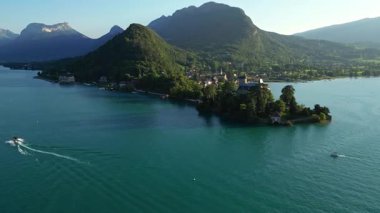 Gün batımında Annecy Gölü 'ndeki Chateau de Duingt' in insansız hava görüntüsü, yemyeşil tepeler ve dağlarla çevrili sakin dağlık sularda süzülen teknelerle. Sinematik altın saat aydınlatması yaratır
