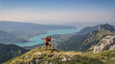 Annecy Gölü ve Fransız Alpleri 'ne tepeden bakan uçurumun kenarında duran genç çift, panoramik yaz dağı ve göl manzarası