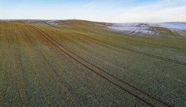 Kışın açık mavi gökyüzü altında kar yamaçları ve traktör lastik izleri olan tarım alanının havadan görünüşü.