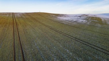 Kışın açık mavi gökyüzü altında kar yamaçları ve traktör lastik izleri olan tarım alanının havadan görünüşü.