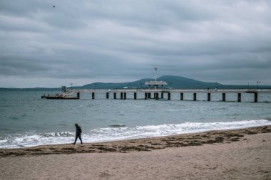Burgaz, Bulgaristan - 9 Kasım 2020: Bulgaristan 'ın Burgaz kentinde boş bir plajda tek başına yürüyen bir kişi. Kış mevsimi, soğuk hava. Bulutlu hava, arka planda deniz köprüsü. Yüksek kalite fotoğraf