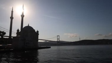 Bir köprü büyük bir binanın önündeki bir su kütlesini kapsar. Gökyüzü bulutlu ve güneş gökyüzünde alçak. Bir kuş sürüsü suyun üzerinde uçuyor.