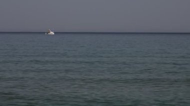Arka planda açık mavi gökyüzü olan bir tekne okyanusta yüzüyor. Sahne sakin ve huzurlu, başka tekne ya da insan yok.