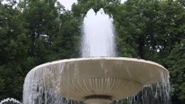Tepesinden su fışkıran büyük beyaz bir çeşme. Su büyük bir kaseye dökülüyor. Çeşme ağaçlarla çevrili.