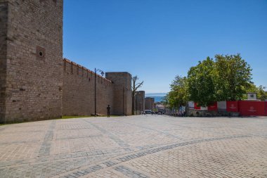 Taş duvar, asfalt kare ve açık gökyüzünün altındaki uzak deniz..