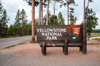 Arka planda bulutlu gökyüzü olan ağaçlara karşı girişteki işaret panosu. Yol üstüne metin ile bilgi işareti. Ünlü Yellowstone Ulusal Parkı 'nın ormanlık manzarası