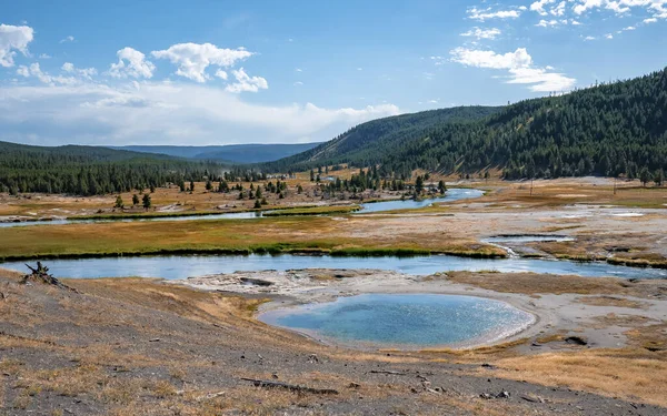 stock image Scenic view of hot spring by river in Midway Geyser Basin. Beautiful geothermal landscape with sky in background during summer. Tourist attraction at famous Yellowstone national park.