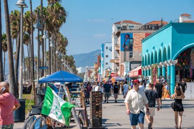 Los Angeles, ABD. 20 Eylül 2022. Erkek ve kadın turistler, Venedik sahilinde arka planda mavi gökyüzü olan binalar ve palmiye ağaçları tarafından kaldırımda yürüyorlar.