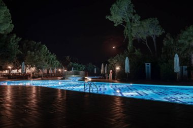 Aydınlatılmış mavi yüzme havuzu üzerinde yaya köprüsü manzarası katlanır şemsiyeler ve ağaçlarla çevrili ve arka planda gökyüzü olan bir turizm beldesi.