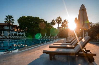 Boş şezlonglar ve katlanmış şemsiyeler güneşli bir günde arka planda parlak güneşle birlikte turizm merkezindeki havuz başında sıralanmış.