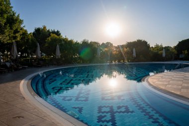 Boş şezlonglar ve şemsiyeler, güneşli bir günde turizm merkezinin arka planında berrak gökyüzü olan kıvrımlı yüzme havuzunun düzenlediği yemyeşil ağaçlarla çevrili.
