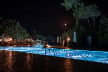 Aydınlatılmış yüzme havuzu üzerindeki yaya köprüsünün manzarası katlanır şemsiyelerle çevrili ve arka planda gökyüzü olan ağaçlarla dolu bir turizm beldesi.