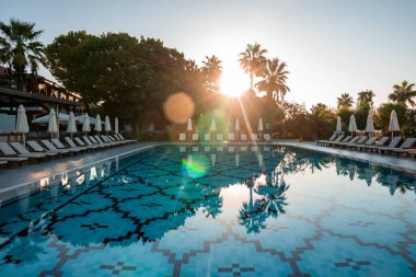 Boş şezlonglar ve katlanmış şemsiyeler, güneşli bir günde turistik bir tatil köyünde yüzme havuzunun etrafında güneşli bir arka planda ve berrak gökyüzü ile düzenlenir.