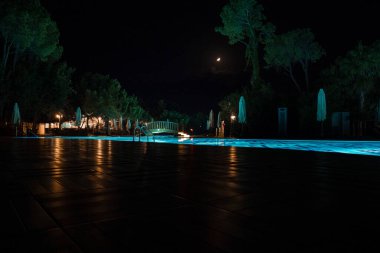 Aydınlatılmış yüzme havuzu manzarası katlanır şemsiyeler ve yemyeşil ağaçlarla çevrili ve geceleri gökyüzü ile birlikte turizm beldesinde büyüyor.